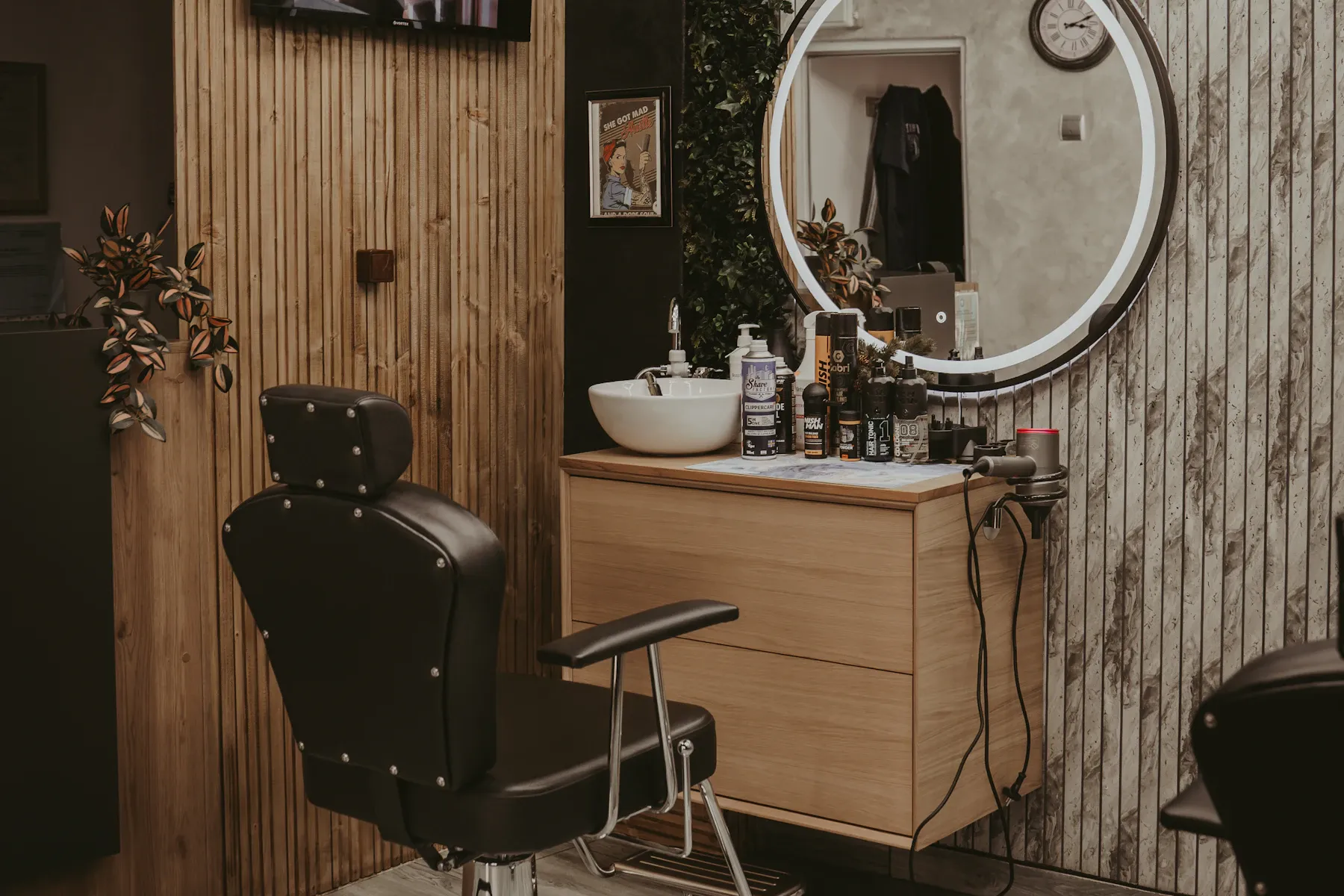 A black styling chair beside a basin, product shelf and lit round mirror in a salon station