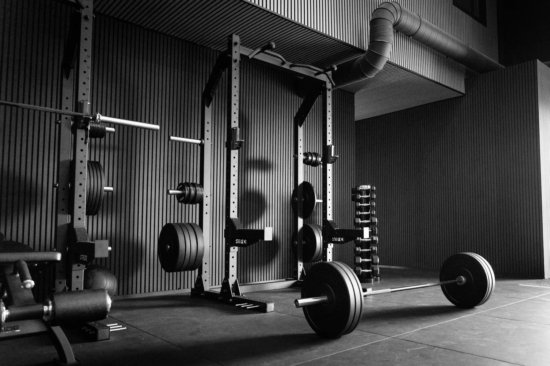 An empty gym with squat racks, a loaded barbell and dumbbells against a dark slatted wall