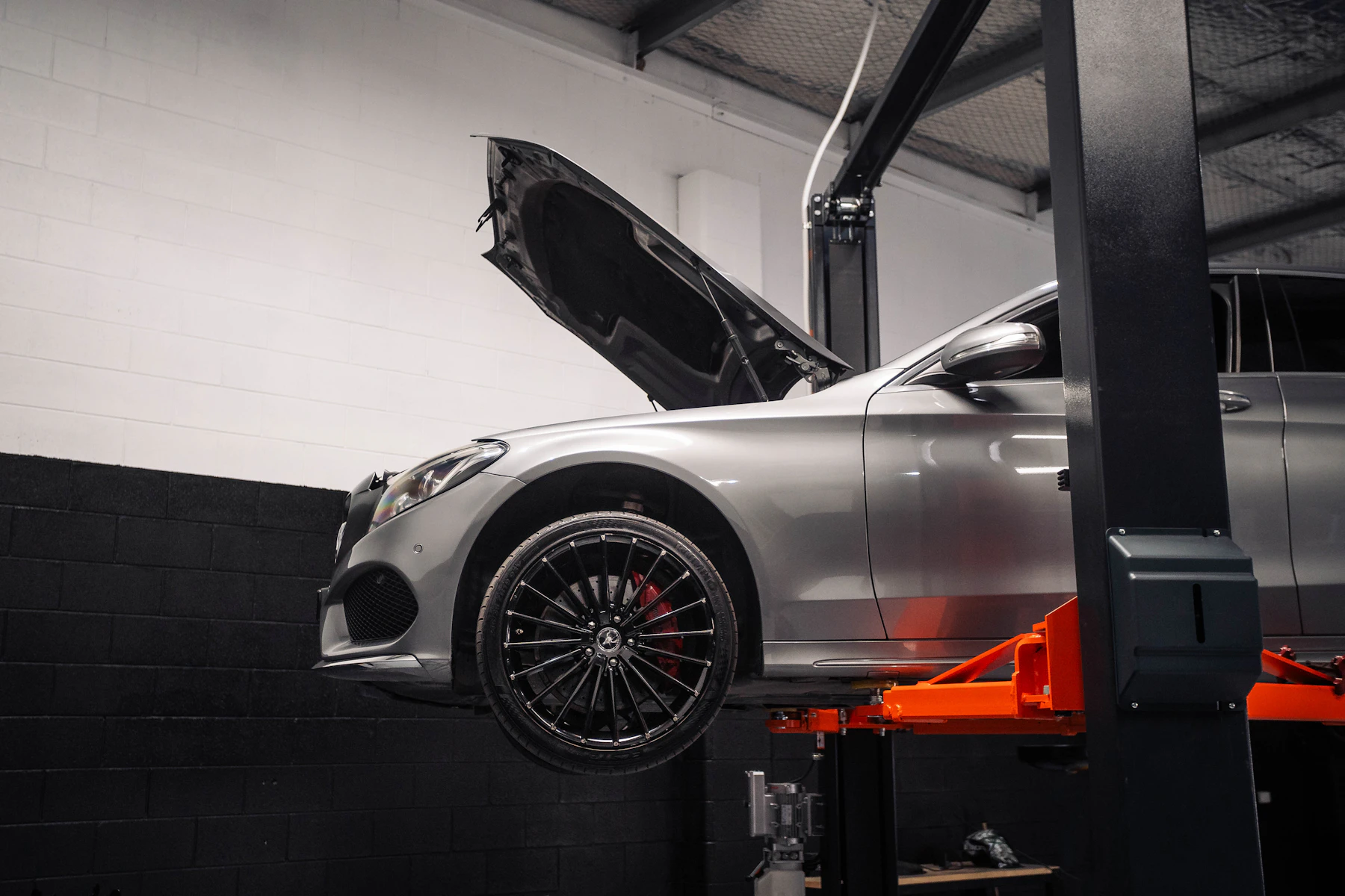 A silver car raised on an orange hydraulic lift in a workshop with its bonnet open